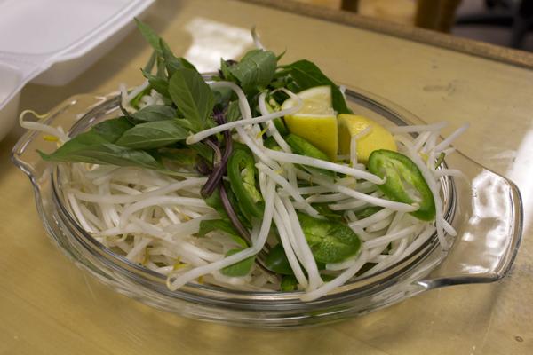 A plate of garnishes to add to the pho includes: lemon, jalapenos, bean sprouts, and mint leaves.
