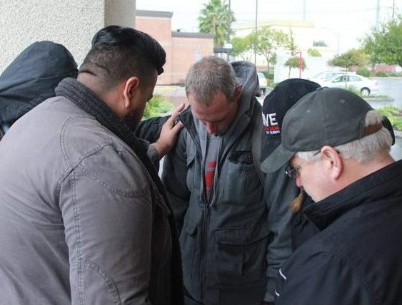 Clockwise, Dawn and Don Nahhas, Providence Church pastor Josh Bueno and Aaron Swan pray with a homeless man to whom they have given a blessing bag of winter essentials on Saturday, Nov. 19. The Nahhas family is working with friends and their church to distribute backpacks to the homeless in memory of their son, who died of liver failure in August. (Photo by John Ferrannini)