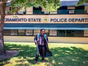 Mulwa James Sumbi poses in front of the Sacramento State Police Department in his graduation robe. Photo courtesy of Jonathan Adorno.
