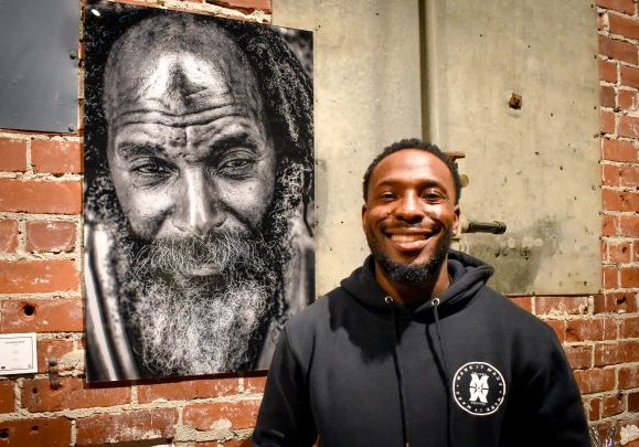 Kachiside Madu at his “Homeless to Heartfull” exhibit on Friday, Nov. 17, 2023 posed in front of his “Black Jesus” black and white portrait at the Brickhouse Gallery. His exhibit features over 8 different black and white pieces, with metal framed portraits, canvas and prints.